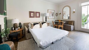 A hotel room with a large bed, a desk, a chair, a sofa, and a dining table. There are framed pictures on the wall, a lamp, and a mirror. - The Chapel | Manor 4BR Charming Charleston (Charleston)