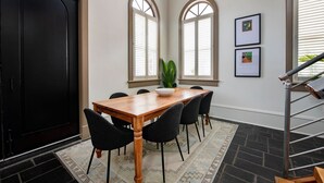 Un coin repas avec une table en bois, des chaises noires, un bol blanc et deux fenêtres équipées de volets.