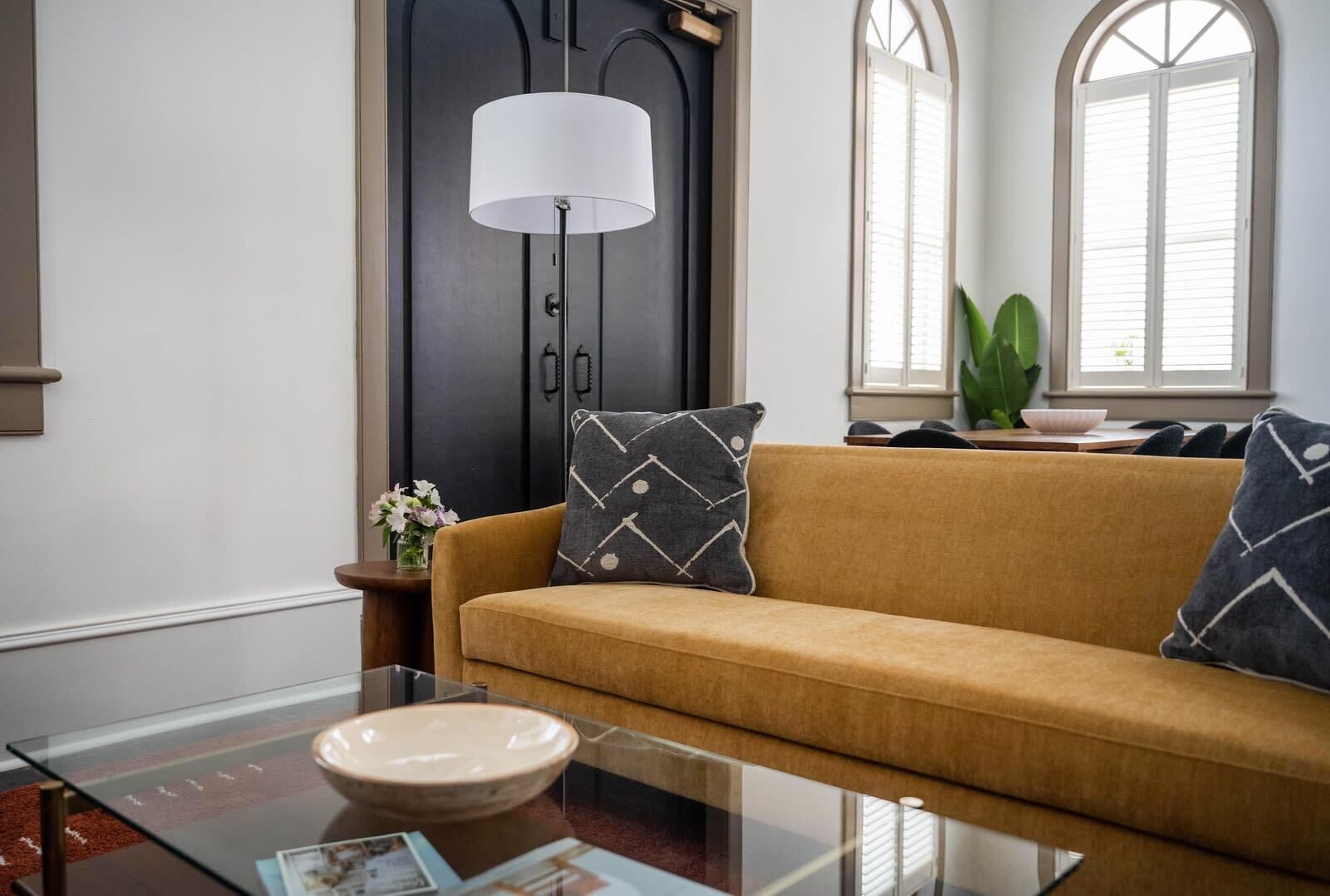 Un salon avec un canapé, une table basse en verre, une lampe et une fenêtre avec des stores.