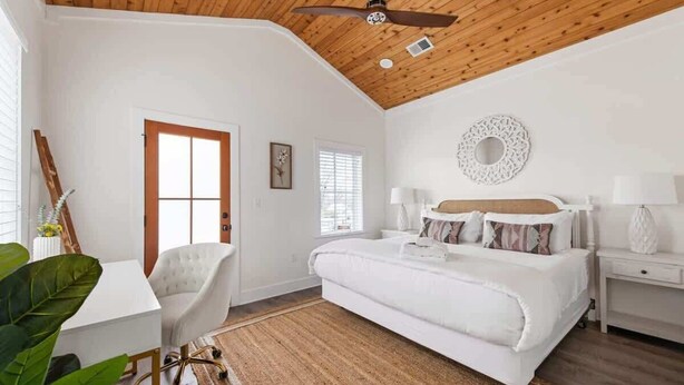 A bedroom with a large bed, a desk, and a chair. There is a ceiling fan, a window, and a decorative wall piece. - Urban Sol | Newly Built in Downtown Charleston! (Charleston)