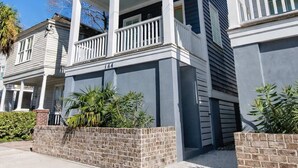 A two-story residential building with a balcony on the upper floor, a brick foundation, and a clear blue sky in the background. - Urban Sol | Newly Built in Downtown Charleston! (Charleston)