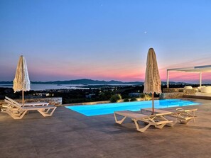 A poolside area with lounge chairs and umbrellas, overlooking a scenic view of a body of water and distant mountains during twilight.