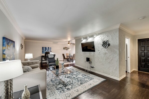 A modern living room with a sofa, coffee table, and decorative items. There is a dining area with chairs and a table in the background.