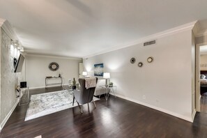 A spacious living room with a dark hardwood floor, a white fireplace, a dining area with a table and chairs, and a large window. - GP66B - |G. Prairie Escape |2BD | Pool | Workspace (Grand Prairie)