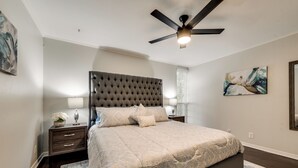 A bedroom with a tufted headboard, a bed with patterned bedding, two bedside tables with lamps, a ceiling fan, and a painting on the wall. - GP66B - Stylish 2BD Condo | Pool | Grand Prairie (Grand Prairie)