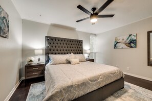 A bedroom with a tufted headboard, a bed with patterned bedding, two bedside tables with lamps, a ceiling fan, and a painting on the wall. - GP66B - Stylish 2BD Condo | Pool | Grand Prairie (Grand Prairie)