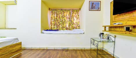 A room with a bed, a window with floral curtains, a small table with a kettle, and a mounted TV.