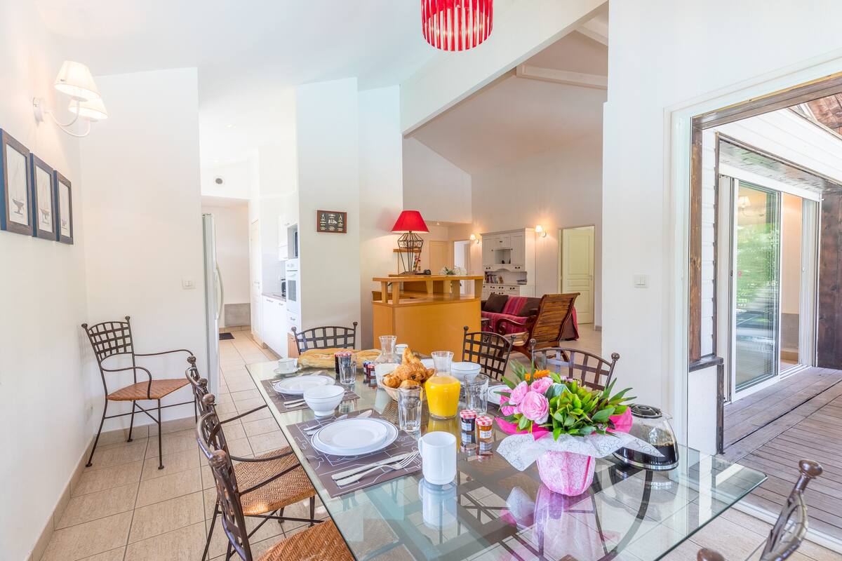 A dining area with a glass table, chairs, and a red lamp. There is a kitchen in the background with cabinets and a countertop.