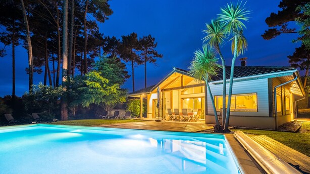 En swimmingpool med liggestole, et hus med terrasse og høje træer i baggrunden.