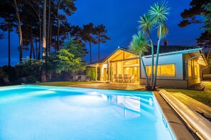 A swimming pool with lounge chairs, a house with a patio, and tall trees in the background. - Madame Vacances Villas Dunes de la Prade (Moliets-et-Maa)