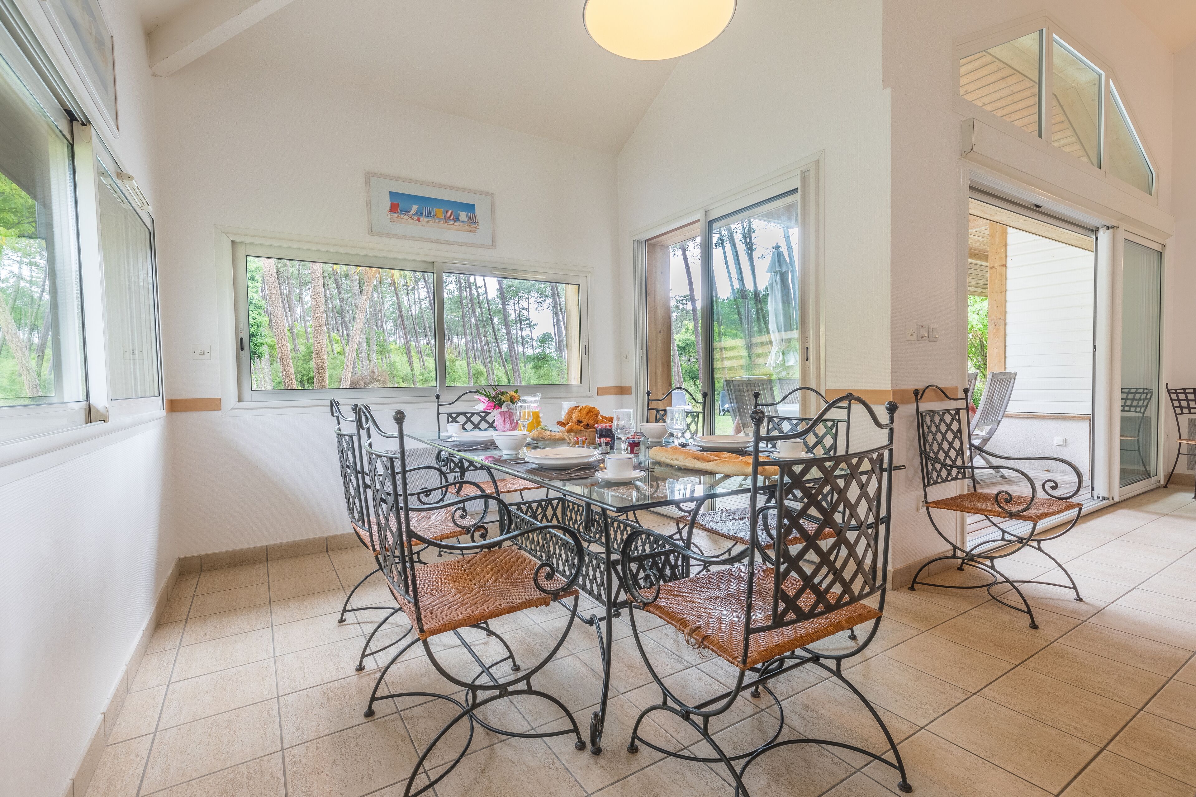 A dining area with a glass table and metal chairs, large windows with a view of trees, and a ceiling light.