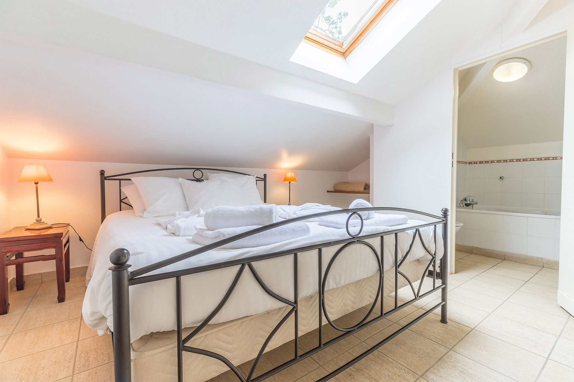 A neatly made bed with white linens and towels, a bedside table with a lamp, and a skylight above.
