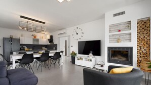 A modern living room with a grey sofa, a dining area with black chairs, a kitchen with white cabinets, a flat-screen TV, and a fireplace. - Stunning home in Labin (Labin)