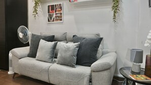 Een grijze bank met meerdere kussens, een staande ventilator, een kleine tafel met een lamp en een plank met decoratieve objecten en planten.
