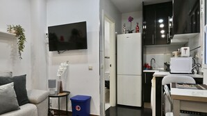Een ruimtelijk ingerichte leefruimte met een bank, een klein bijzettafeltje, een blauwe prullenbak, een aan de muur bevestigde televisie, een koelkast, een magnetron, en een eethoek met een stoel en tafel.