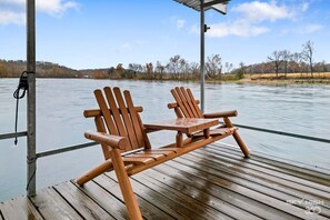 Eine Holzterrasse mit zwei Adirondack-Stühlen und einem Tisch, mit Blick auf ruhiges Wasser und Bäume im Hintergrund.