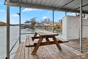 A wooden picnic table under a metal roof overlooking a river with houses in the background. - Riverfront Relaxation (Cotter)
