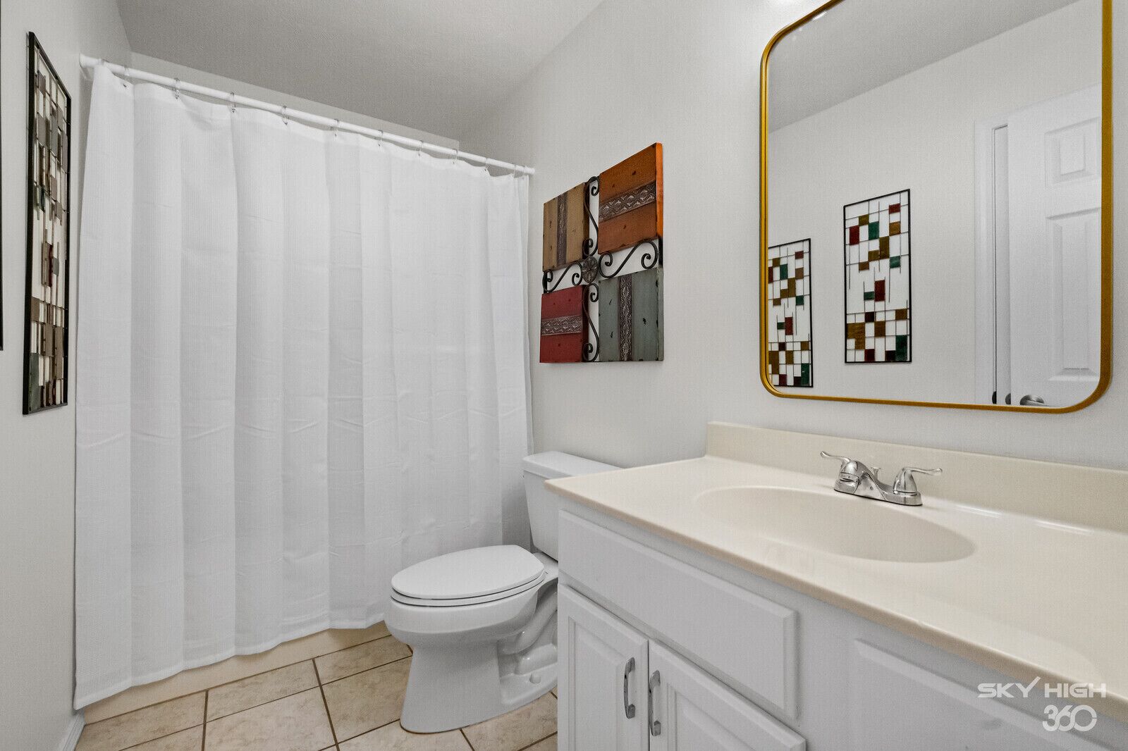 A bathroom with a white toilet, a white sink, and a shower curtain. There is a mirror with a gold frame and a wall-mounted artwork above the sink.