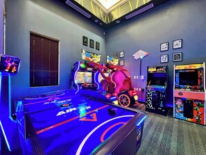 Une salle avec une table de billard, des jeux d’arcade et un espace de jeux éclairé par des néons.