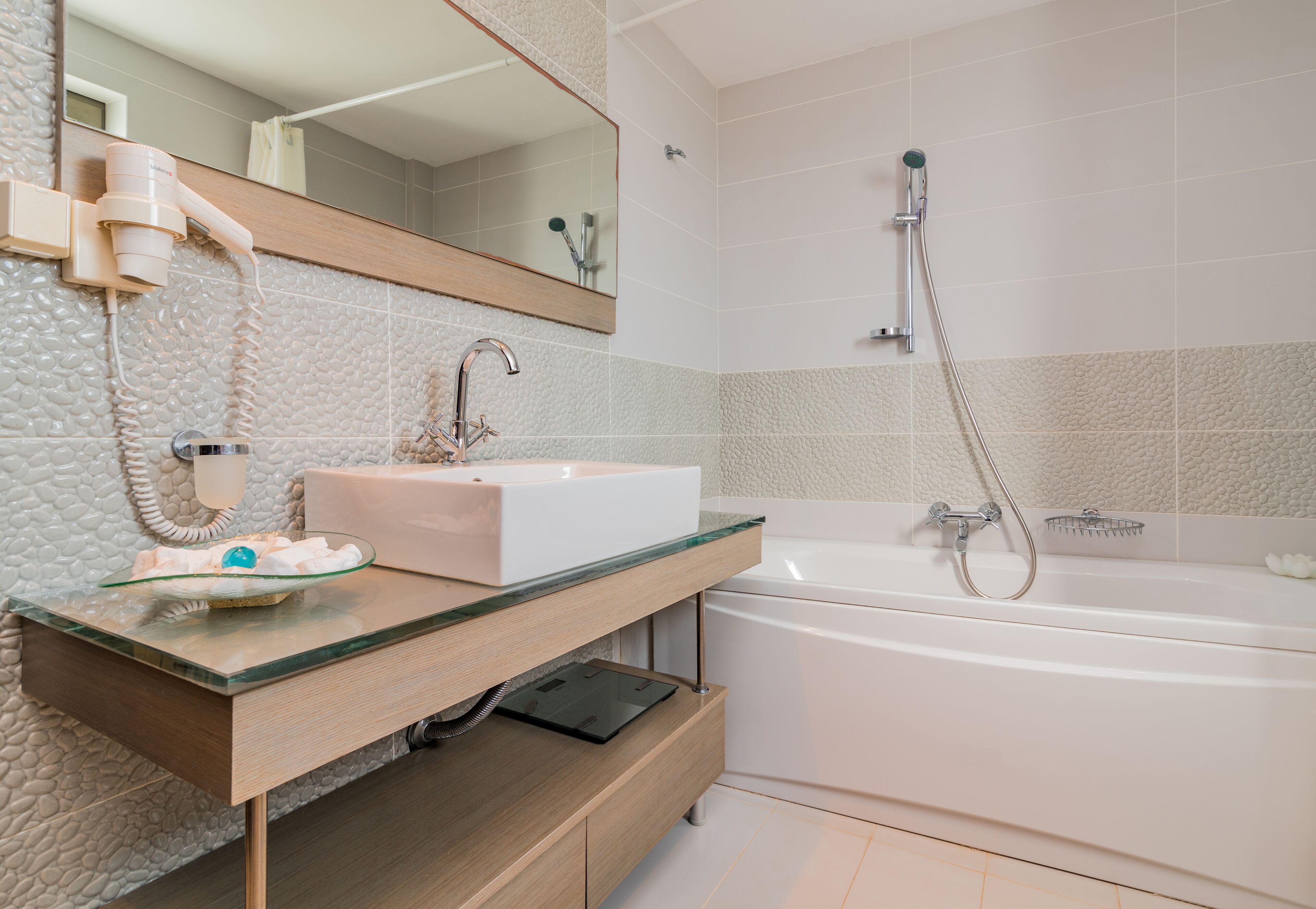 Un baño moderno con encimera de vidrio, un lavabo blanco, espejo integrado, cabezal de ducha manual y bañera.