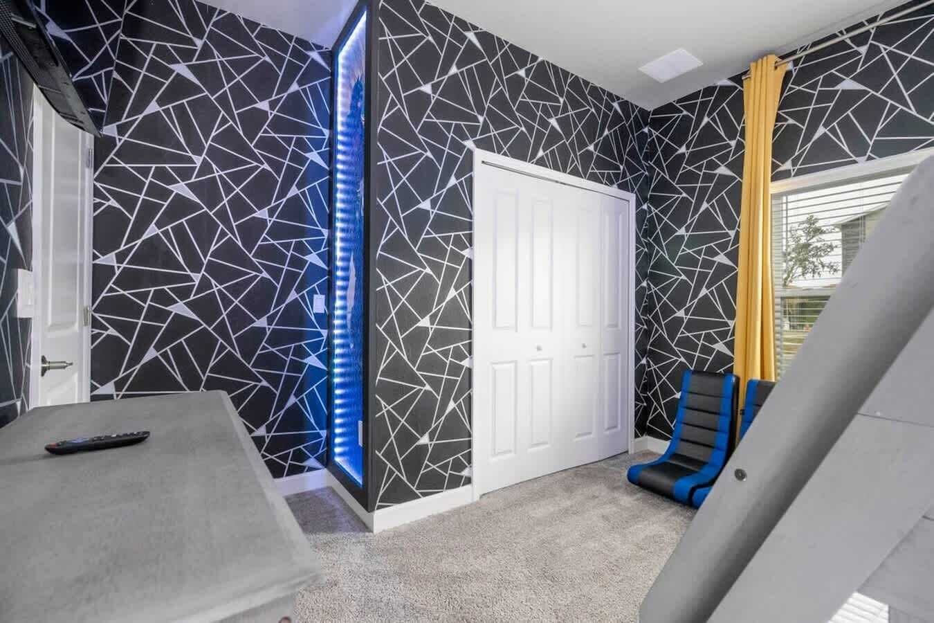 A room with a geometric patterned wall, a white door, a blue chair, and a remote control on a table.