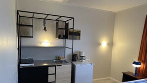 Et kompakt hotelværelse med tekøkken, en seng med blå og hvidstribet sengetæppe, et natbord med lampe og en vægmonteret hylde med vinglas.