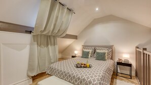 A bedroom with a sloped ceiling, a bed with patterned bedding, bedside tables with lamps, and a bench. - Charmant Duplex 30 min Paris (Corbeil-Essonnes)