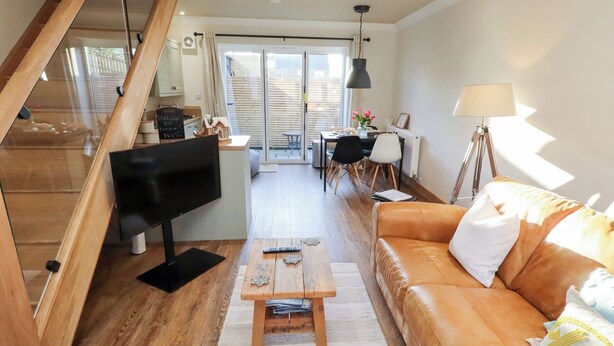 Ein modernes Wohnzimmer mit einer Holztreppe, einem Fernseher, einem Holztisch, einem Ledersofa und einem Essbereich mit Tisch und Stühlen.
