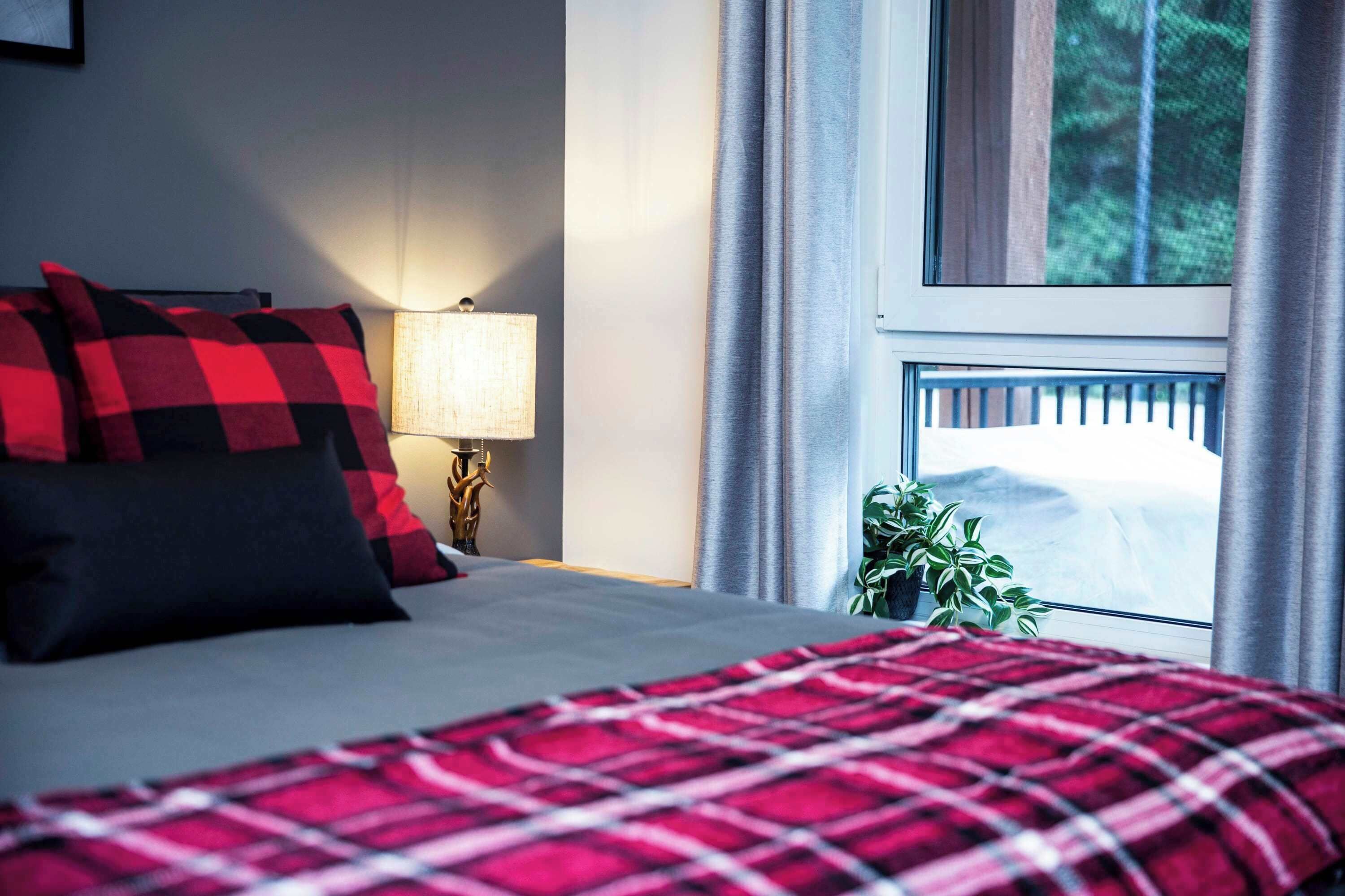 A bed with a red and black checkered blanket, a bedside lamp, a window with curtains, and a plant on the windowsill.