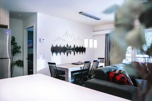 A modern living room with a dining area, a sofa, and a wall-mounted artwork of mountains. - Absolutely Revy - Ground Level Condo with Hot Tub (Revelstoke)