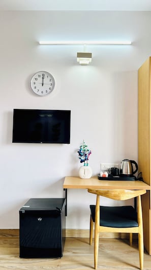 A small, modern room with a wooden table, a black chair, a TV mounted on the wall, a clock, and a small plant.