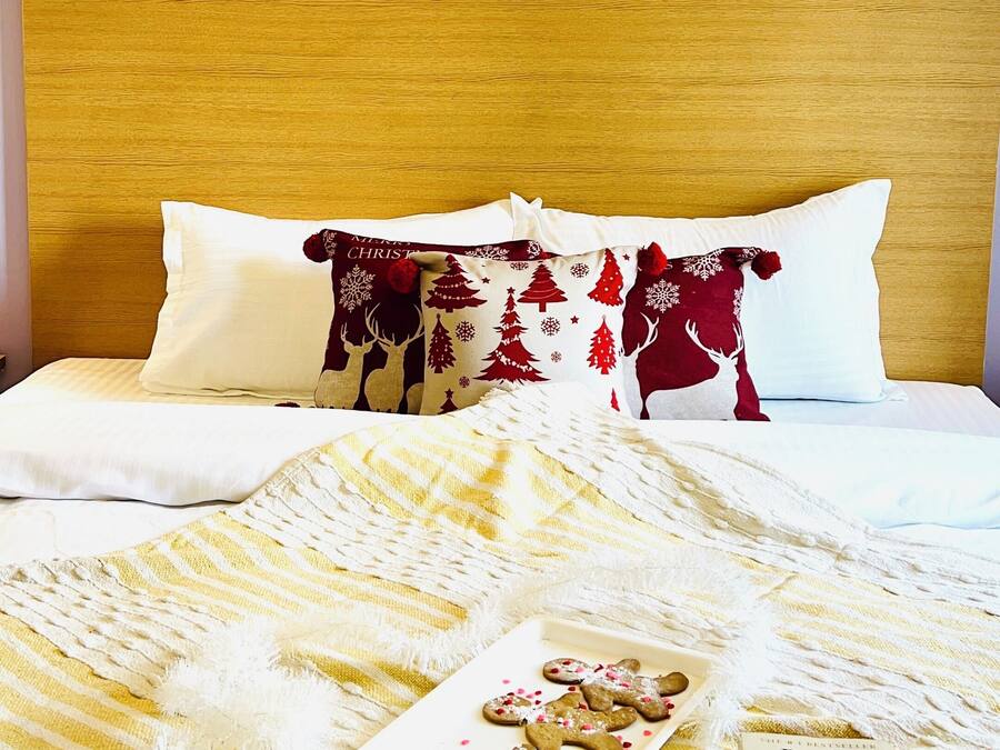 A neatly made bed with a white and maroon patterned pillow, a yellow and white striped blanket, and a tray with two mugs of coffee and a book titled "Adultery" by Paulo Coelho.