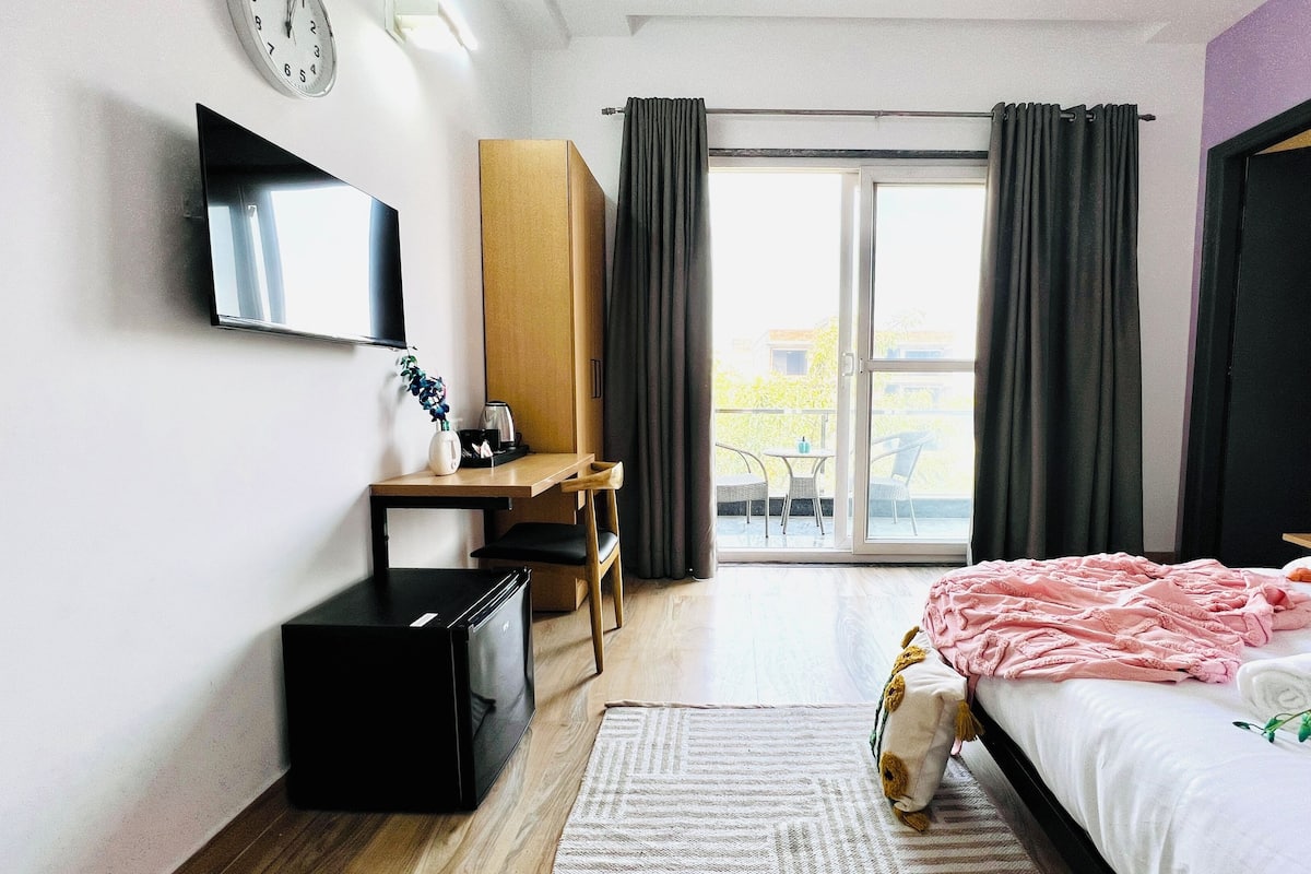 A modern hotel room with a bed, a desk, a TV, and a balcony with curtains.