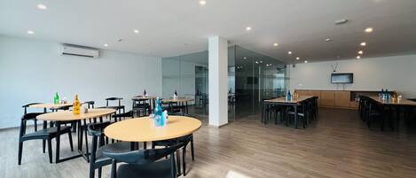 A spacious conference room with multiple tables and chairs, a flat-screen TV mounted on the wall, and a glass partition separating the dining area from the meeting space.