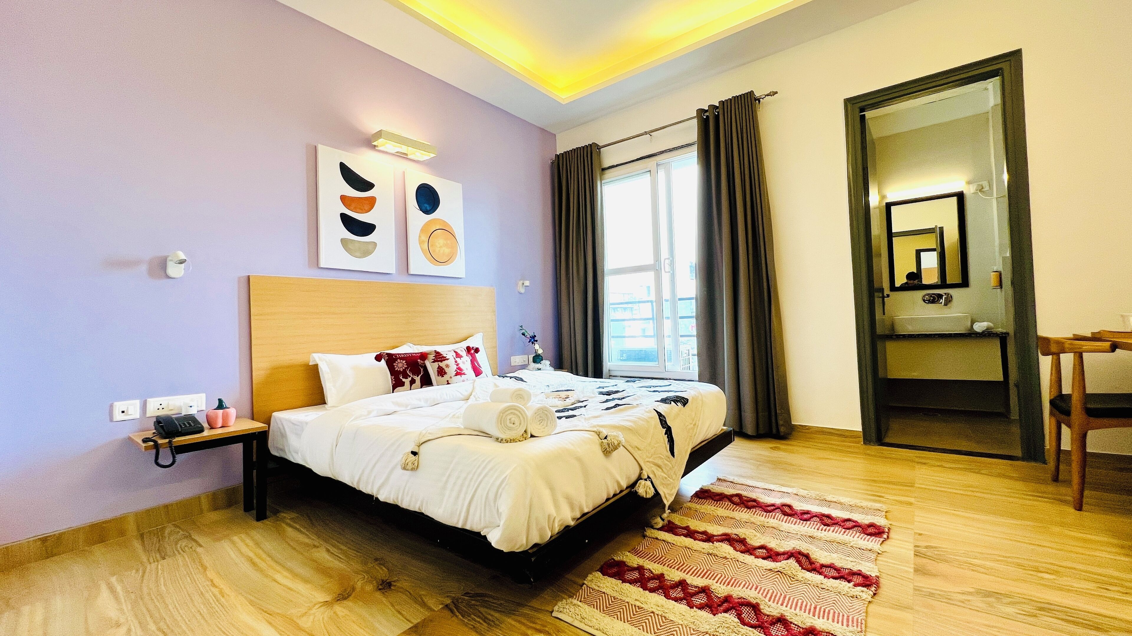 Ein Hotelzimmer mit Bett, Nachttisch, Lampe, Kunstwerk, Fenster mit Vorhängen und Spiegel.