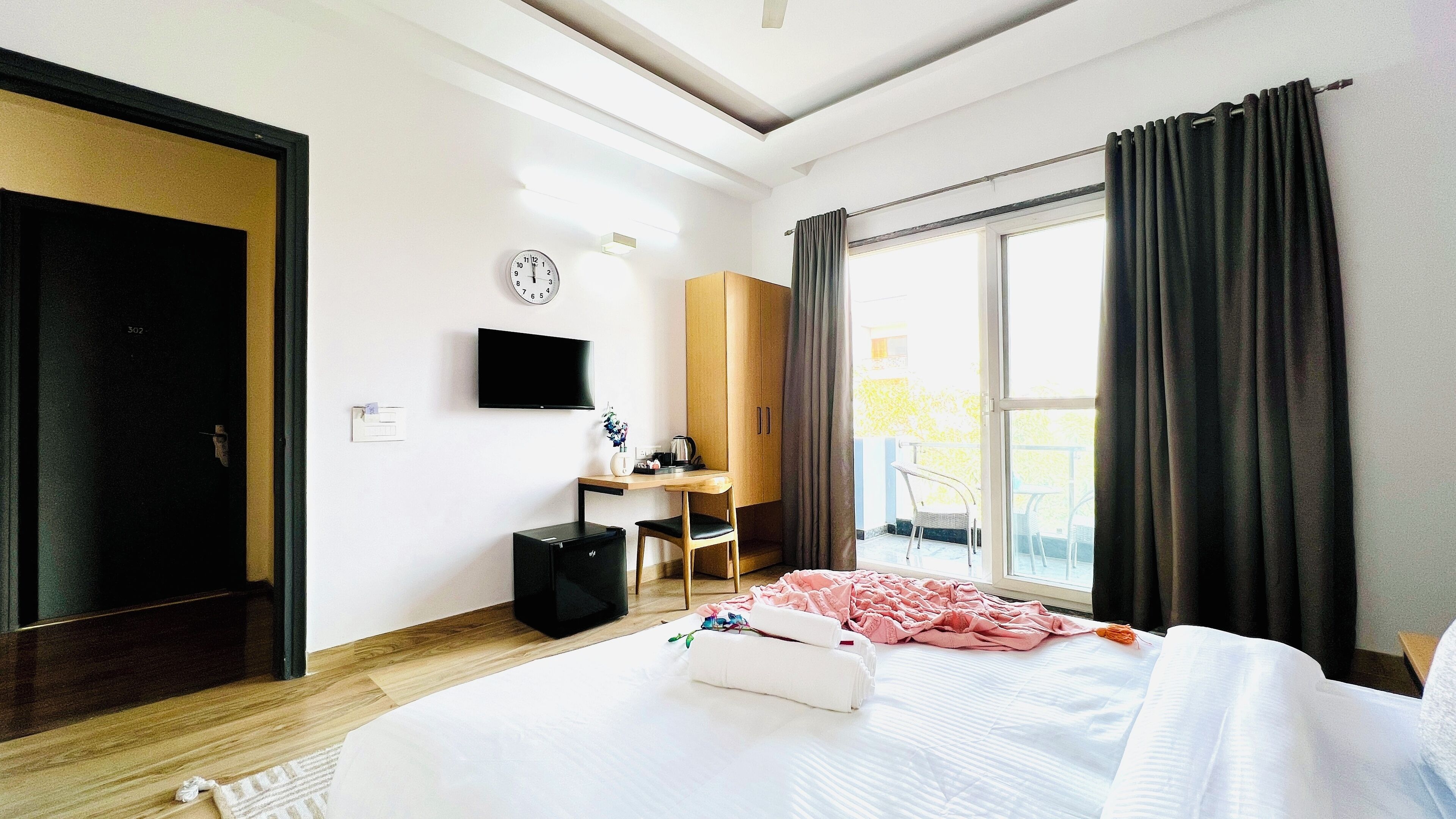 A hotel room with a bed, a desk with a chair, a TV, a wardrobe, a clock, and a window with curtains.