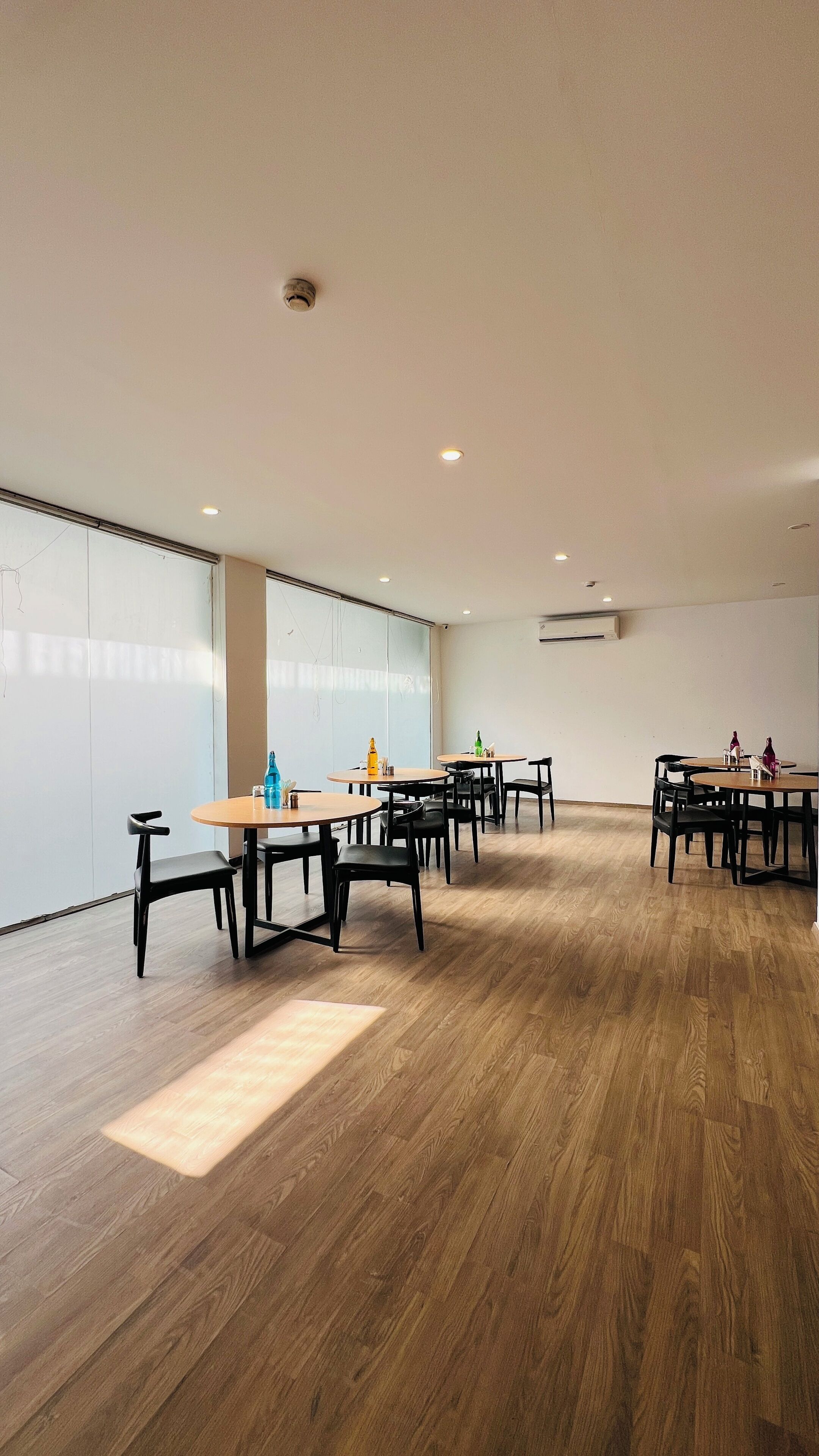 A spacious room with multiple round tables and chairs, large windows, and a wooden floor.