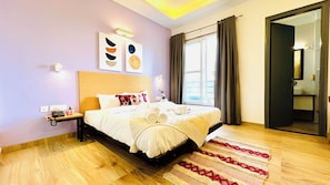 A hotel room with a wooden floor, a bed with white bedding, a bedside table with a phone, and a window with curtains.