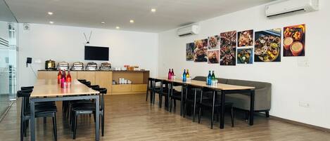 Un comedor moderno con mesas y sillas de madera, una zona de cocina con electrodomésticos y una pared decorada con motivos gastronómicos.