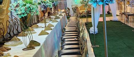 A long banquet table with a white tablecloth, surrounded by chairs and decorated with floral arrangements.