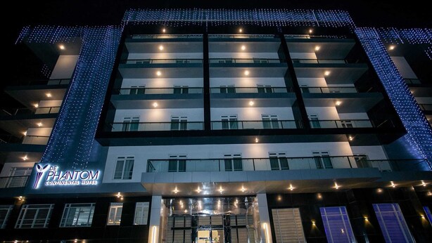 Un hôtel à plusieurs étages, doté d’une façade moderne illuminée de lumières bleues, avec une entrée imposante marquée par une porte en verre et une enseigne bien visible indiquant « De Phantom Continental Hotel ».