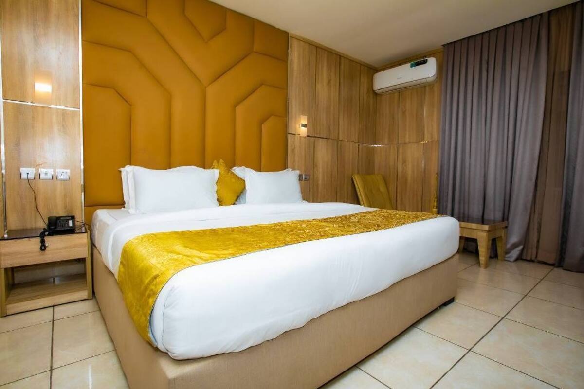 Quarto de hotel com uma cama grande, cabeceira de madeira, mesa de cabeceira com abajur e unidade de ar condicionado.