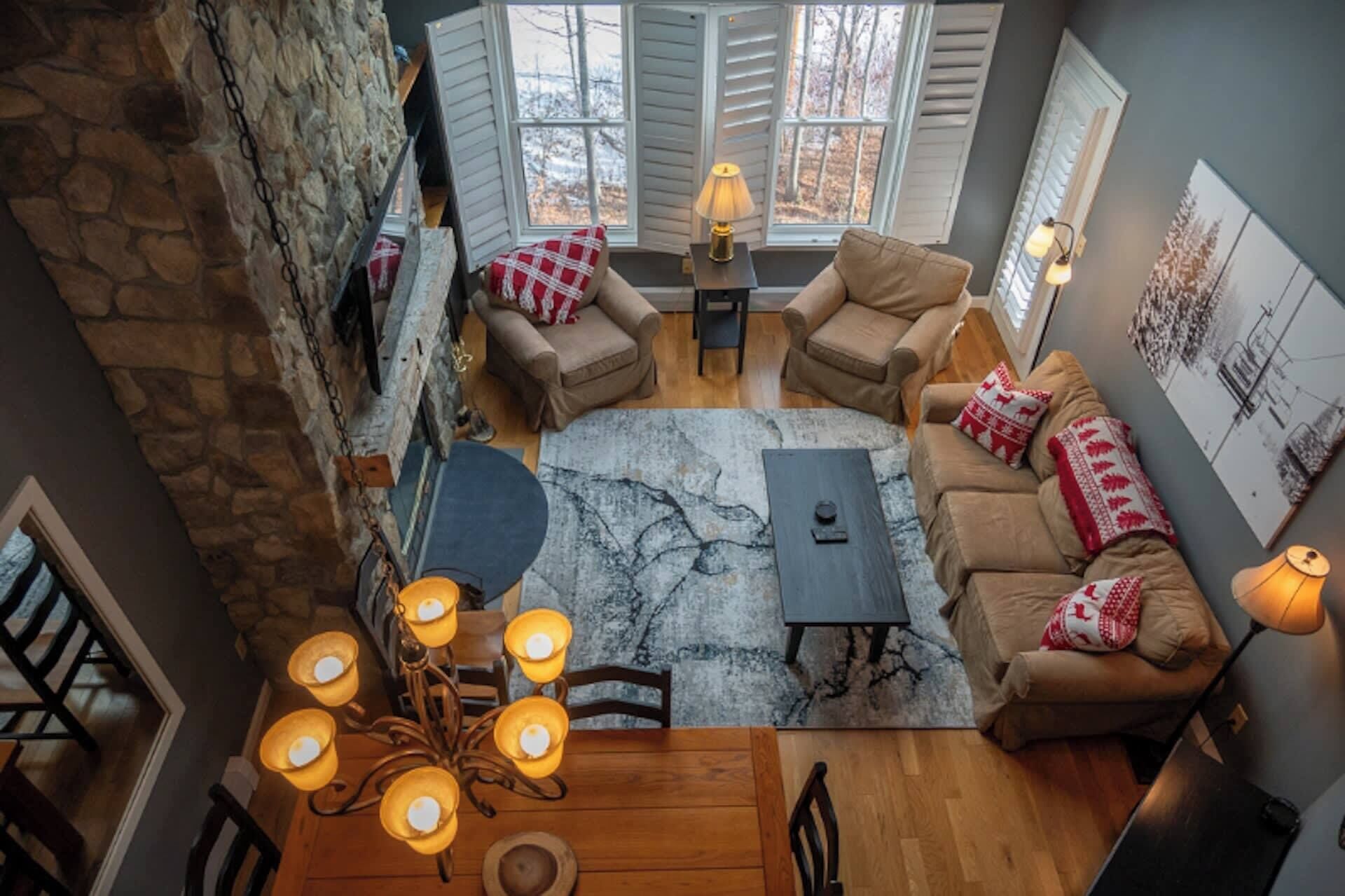 A cozy living room with a stone wall, a fireplace, a chandelier, and a view of the outdoors through large windows.