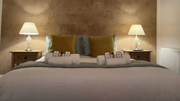 A neatly made bed with two pillows, two bedside tables with lamps, and a wall with a textured finish. - St Cuthbert St Royal Suites (Kirkcudbright)