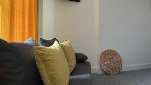 A living room with a grey sofa, yellow and grey cushions, a round textured wall decoration, and a sign above reading "R suites". - St Cuthbert St Royal Suites (Kirkcudbright)