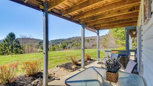 A covered patio with a glass table and chairs, a stone pathway, and a view of a green field and hills.
