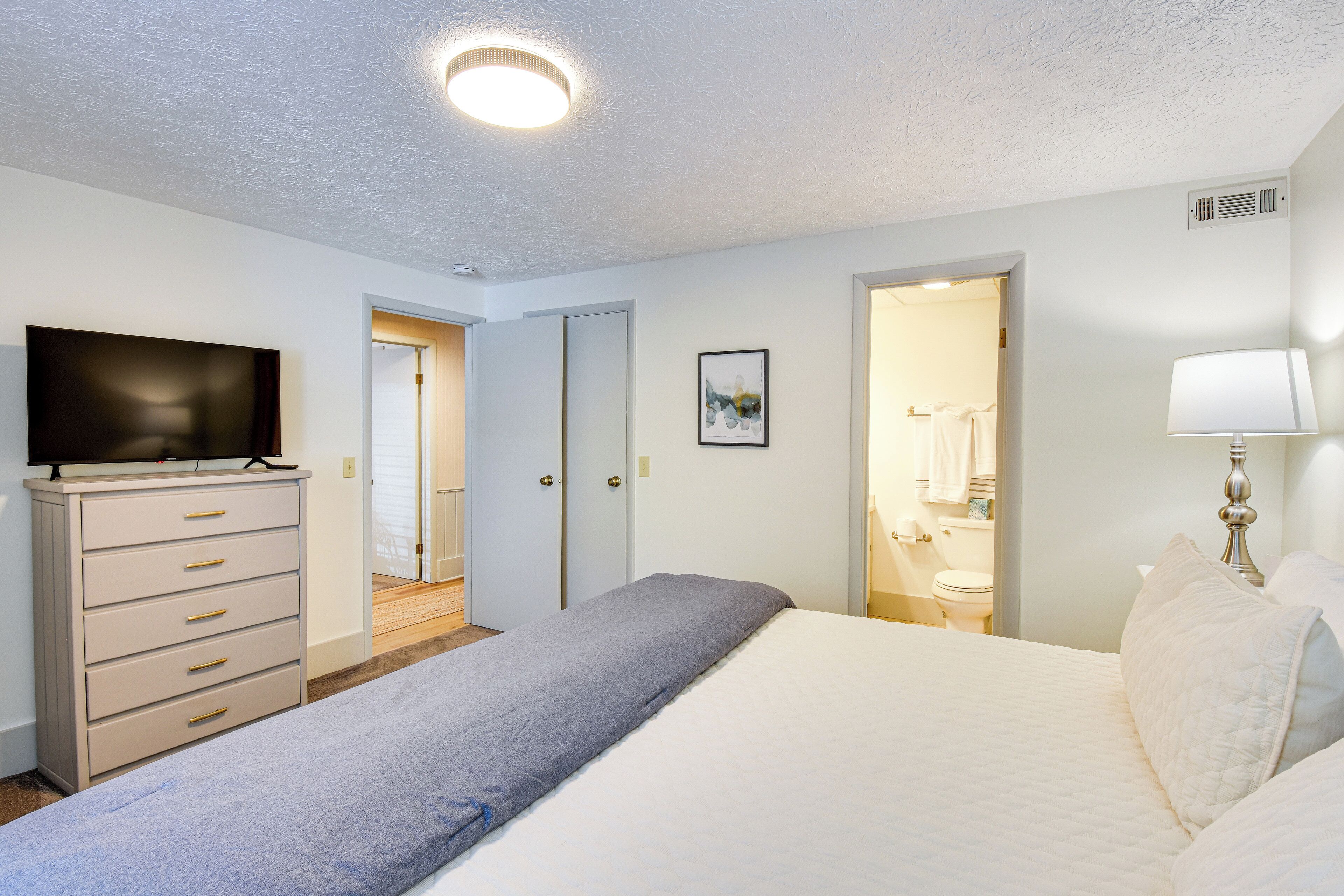 Un dormitorio con cama, cómoda, televisión y baño visible a través de una puerta abierta.