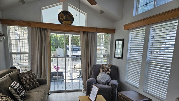A living room with a grey sofa, a wooden table with a menu, a grey armchair with a leopard print pillow, and a view of a backyard through large windows.