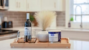 A wooden tray with a bottle of wine, two glasses, a vase with white flowers, and a container of lavender candle wax.