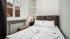 A neatly made bed with white linens and rolled towels, a bedside table with a lamp and vase of flowers, a radiator, and a window with a view of buildings. - Apartamenty Sun & Snow Przy Barbakanie (Warsaw)
